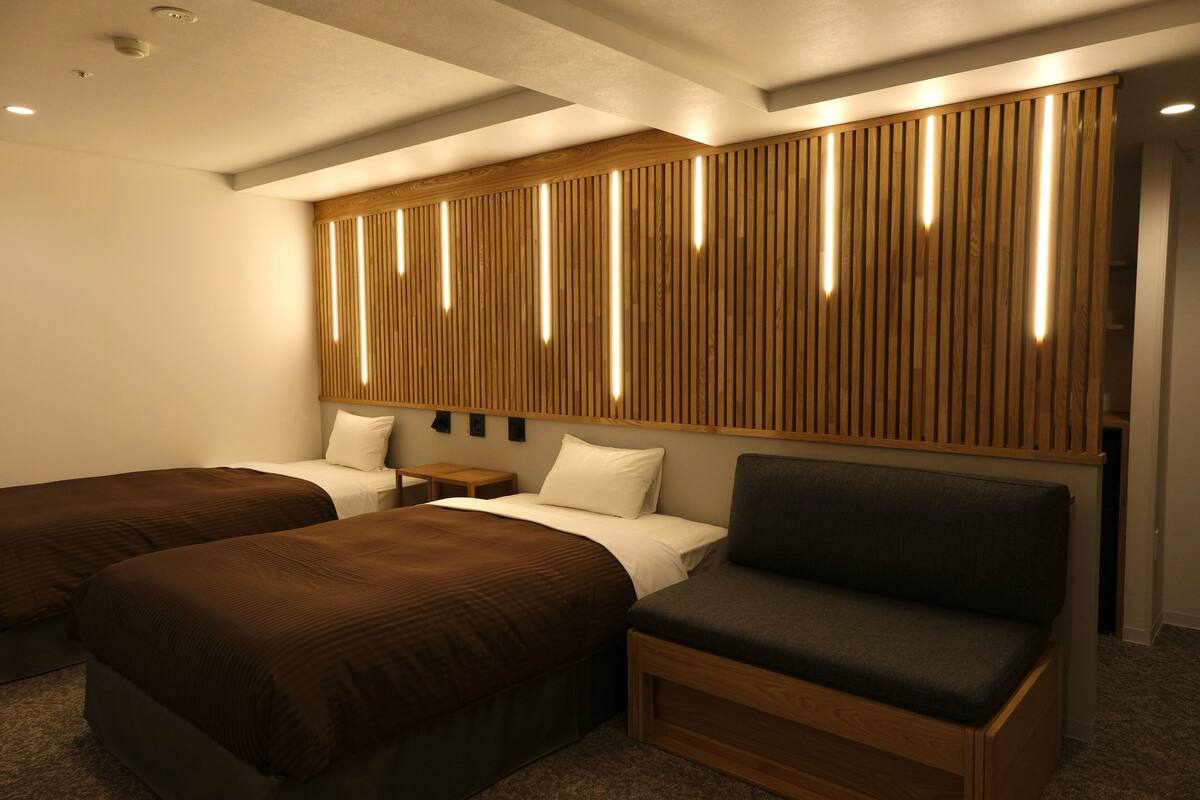 A hotel room with two single beds, a sofa, a wooden headboard with vertical slats, and a small bedside table.