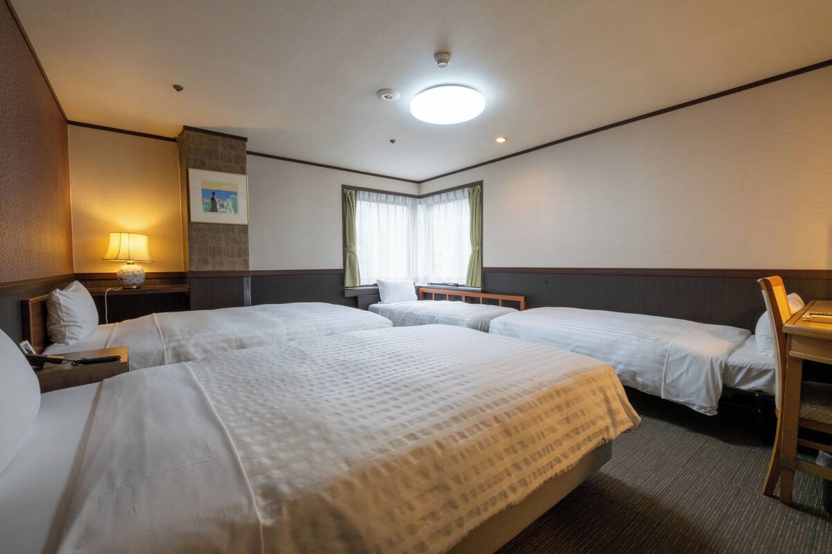 A hotel room with two beds, a desk, a chair, a lamp, a window with curtains, and a ceiling light.