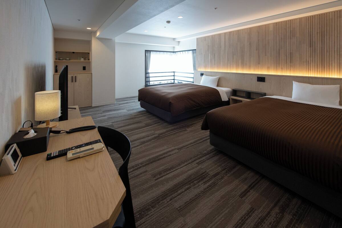 A hotel room with a bed, bedside table, lamp, and a desk with a phone and remote controls.