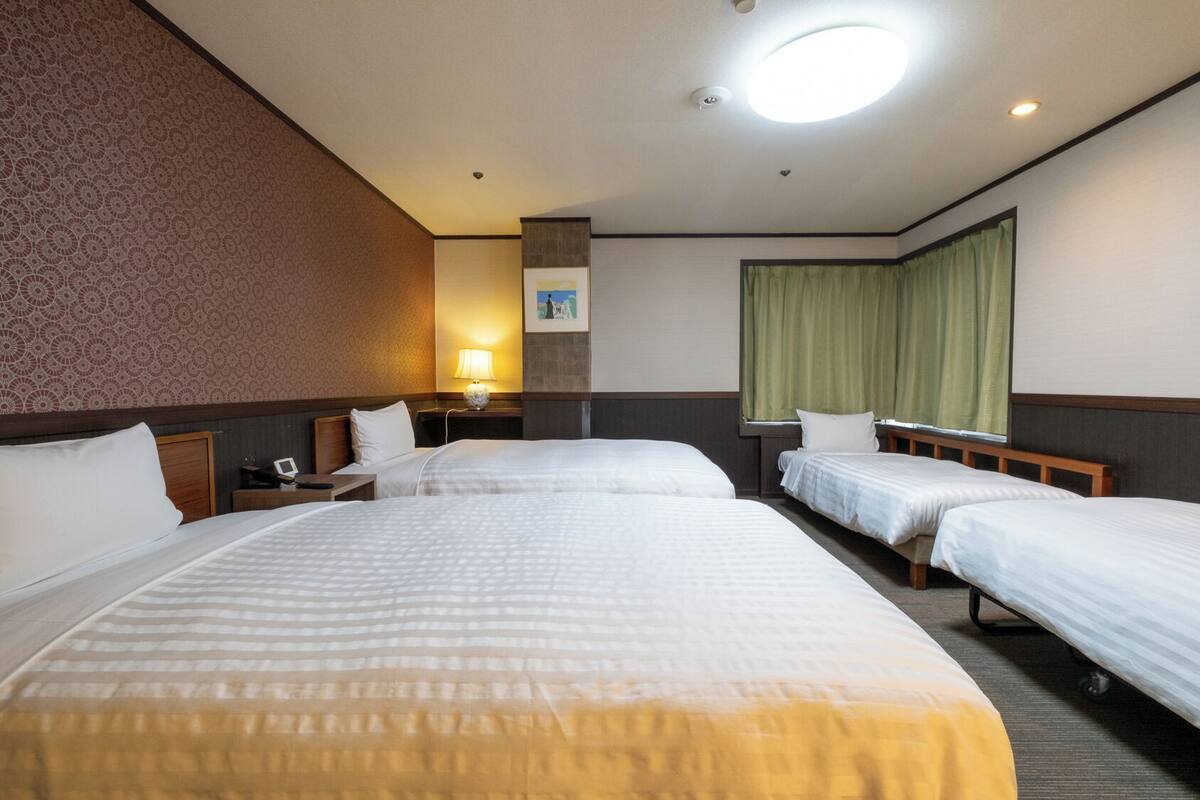 A hotel room with three single beds, a desk with a lamp, a small table, and a window with curtains.