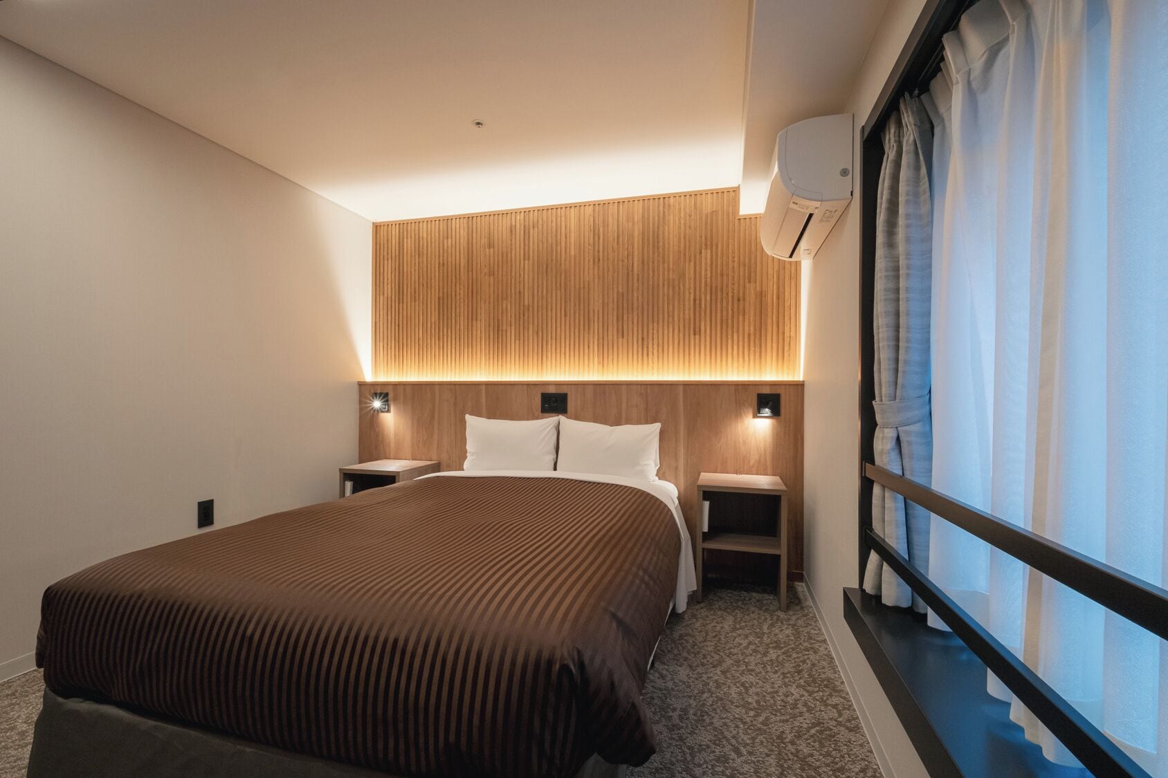 A hotel room with a large bed, wooden headboard, bedside lamps, a window with curtains, and an air conditioning unit.
