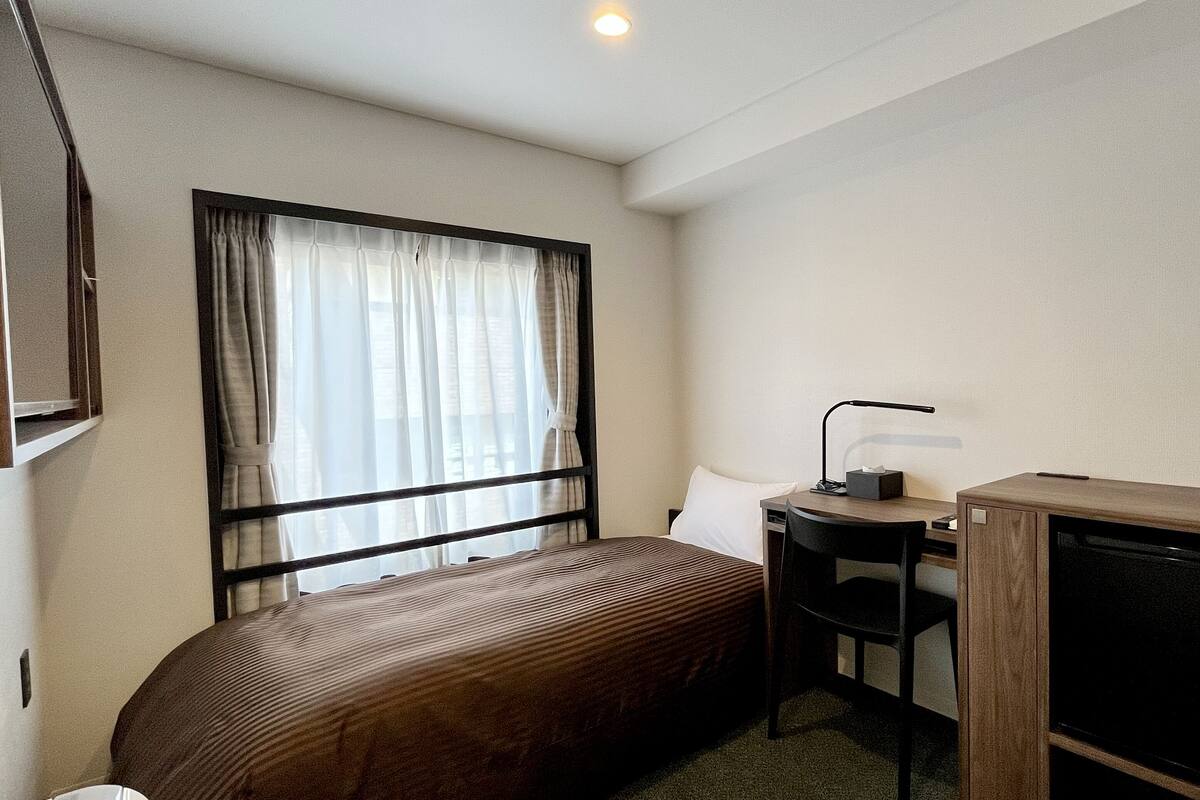 A small hotel room with a single bed, a desk with a chair, a TV, and a window with curtains.
