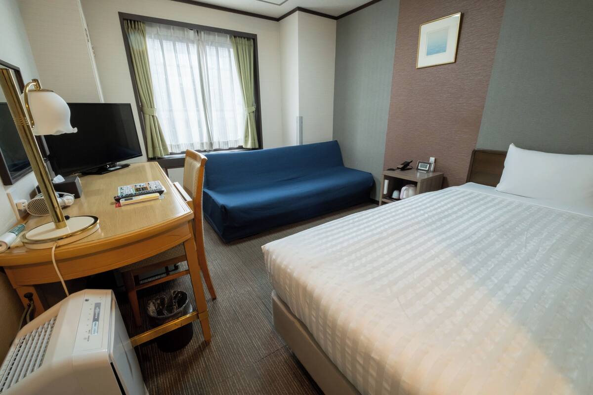 A hotel room with a bed, a desk with a computer, a television, a sofa, and a window with curtains.