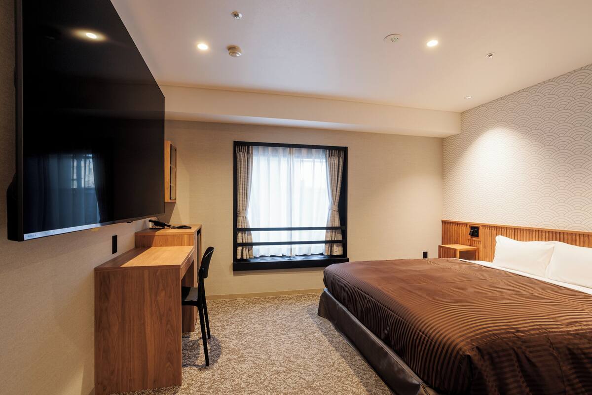 A hotel room with a bed, a desk, a TV, a window with curtains, and a patterned wall.