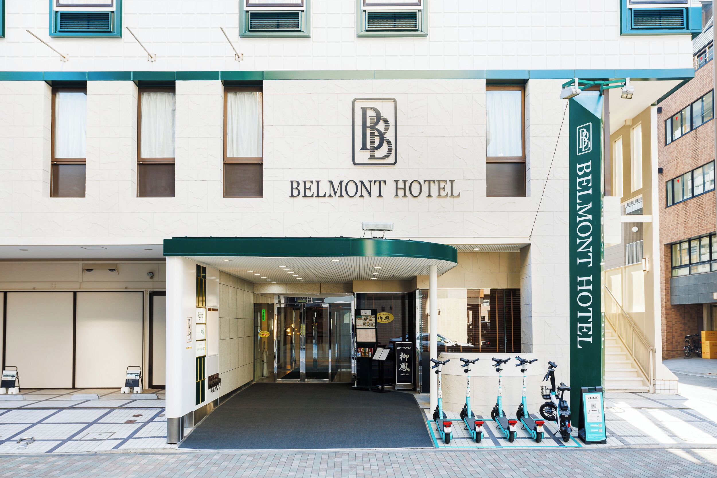 La façade de l’hôtel Belmont, avec une entrée bien visible, une enseigne et des trottinettes électriques garées à l’extérieur.