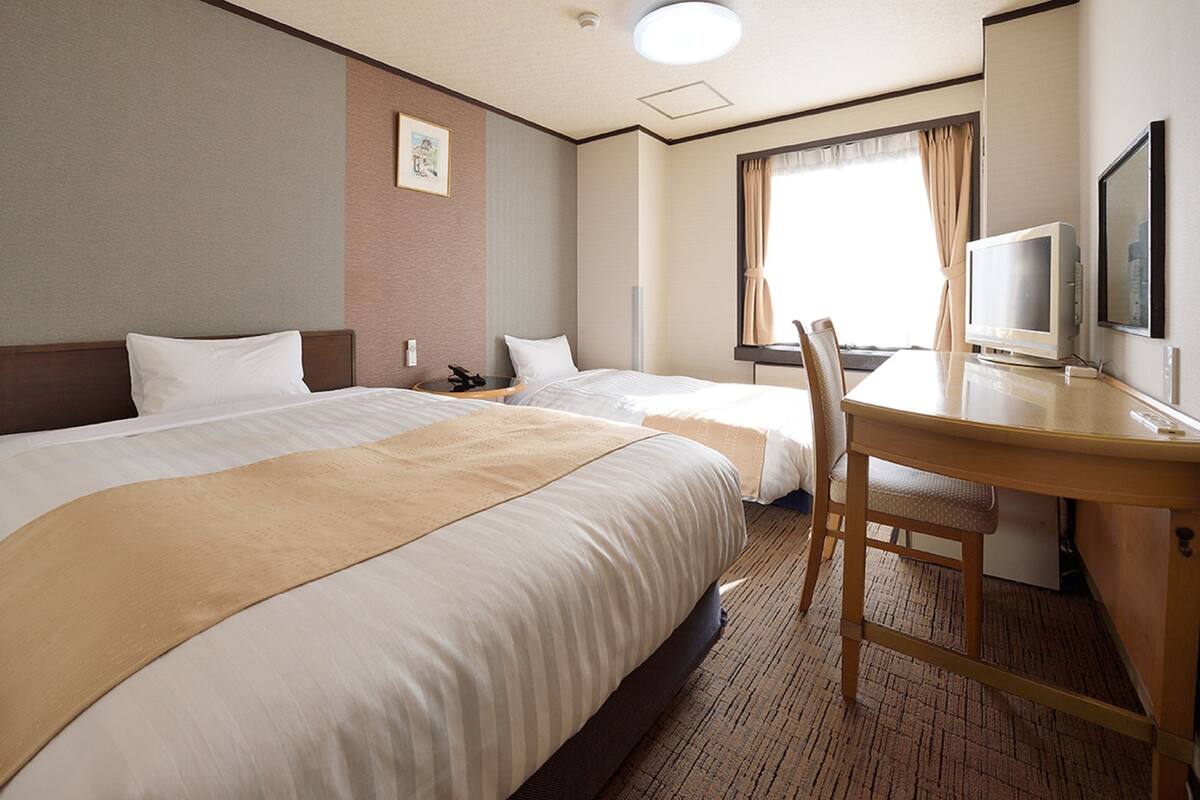 A hotel room with two beds, a desk with a chair, a television, and a window with curtains.