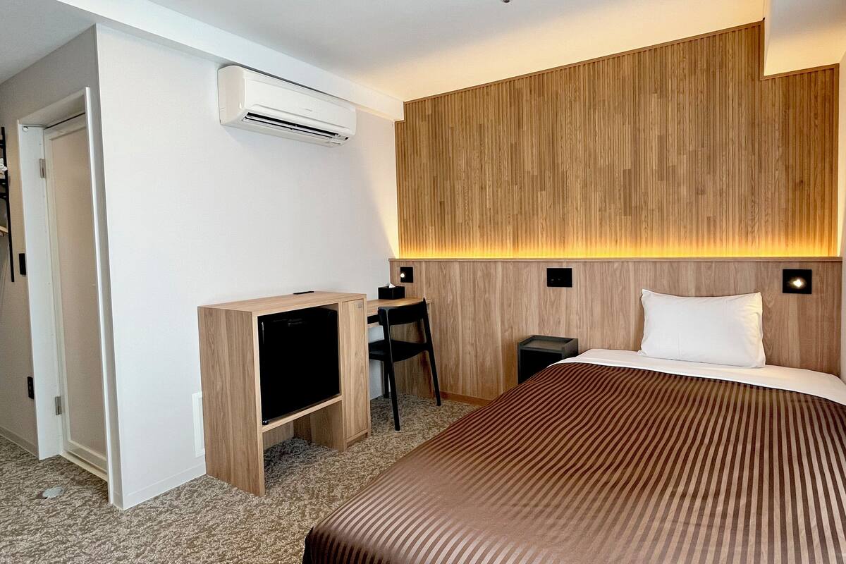 A hotel room with a bed, a desk with a chair, a television, and an air conditioning unit.