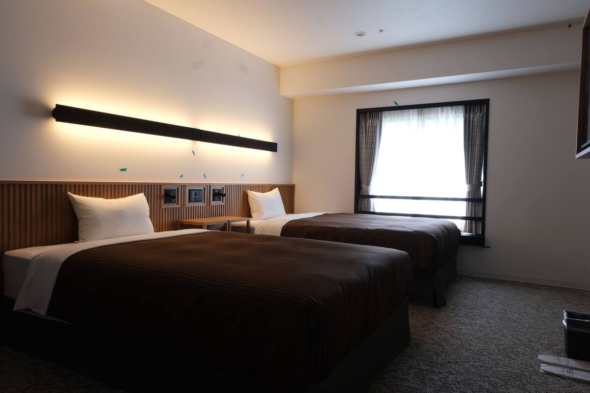 A hotel room with two beds, a large window with curtains, a wall-mounted light fixture, and a flat-screen TV.