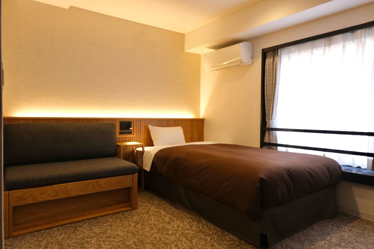 A hotel room with a bed, a sofa, a bedside table, and a window with curtains.