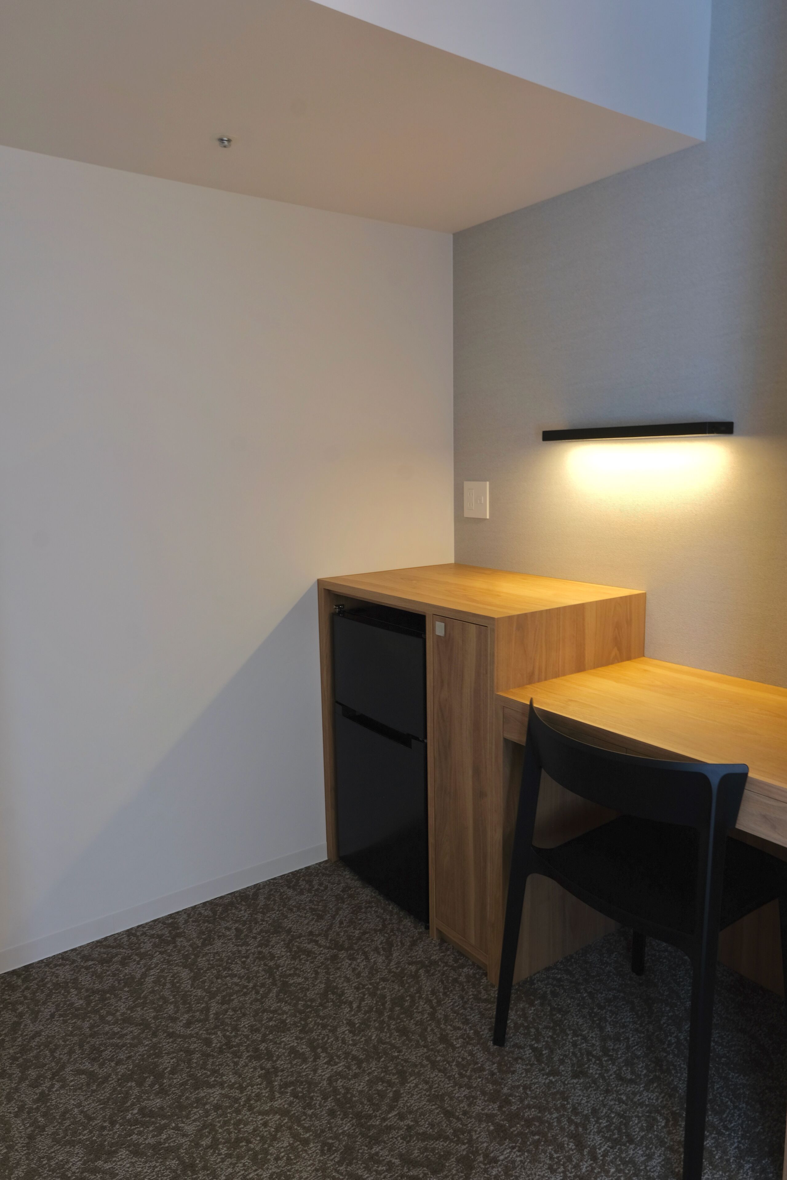 A corner room with a wooden desk, a black chair, and a wall-mounted shelf.