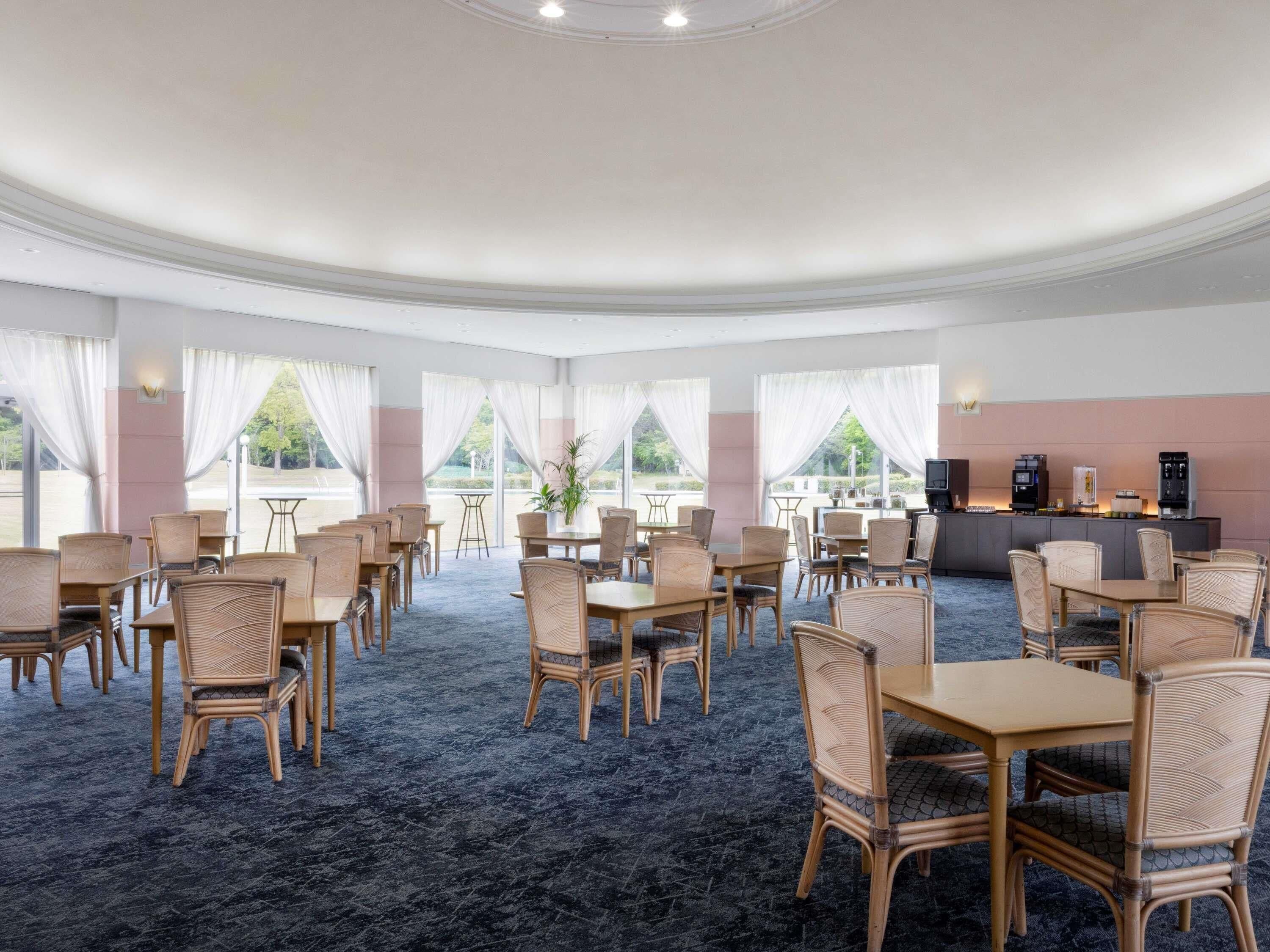 A hotel room with a curved ceiling, large windows with white curtains, wicker furniture, and a minibar.