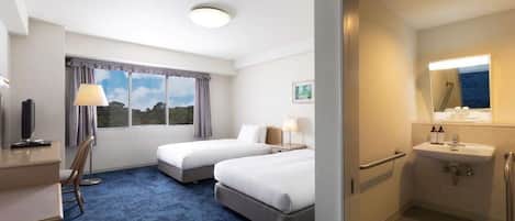 Habitación de hotel con dos camas, un escritorio, una silla, una lámpara de escritorio, un ventanal con vistas y un baño con lavamanos y espejo.