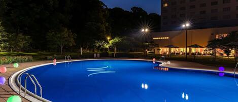 A swimming pool with a tiled deck, surrounded by trees and a building with illuminated windows.
