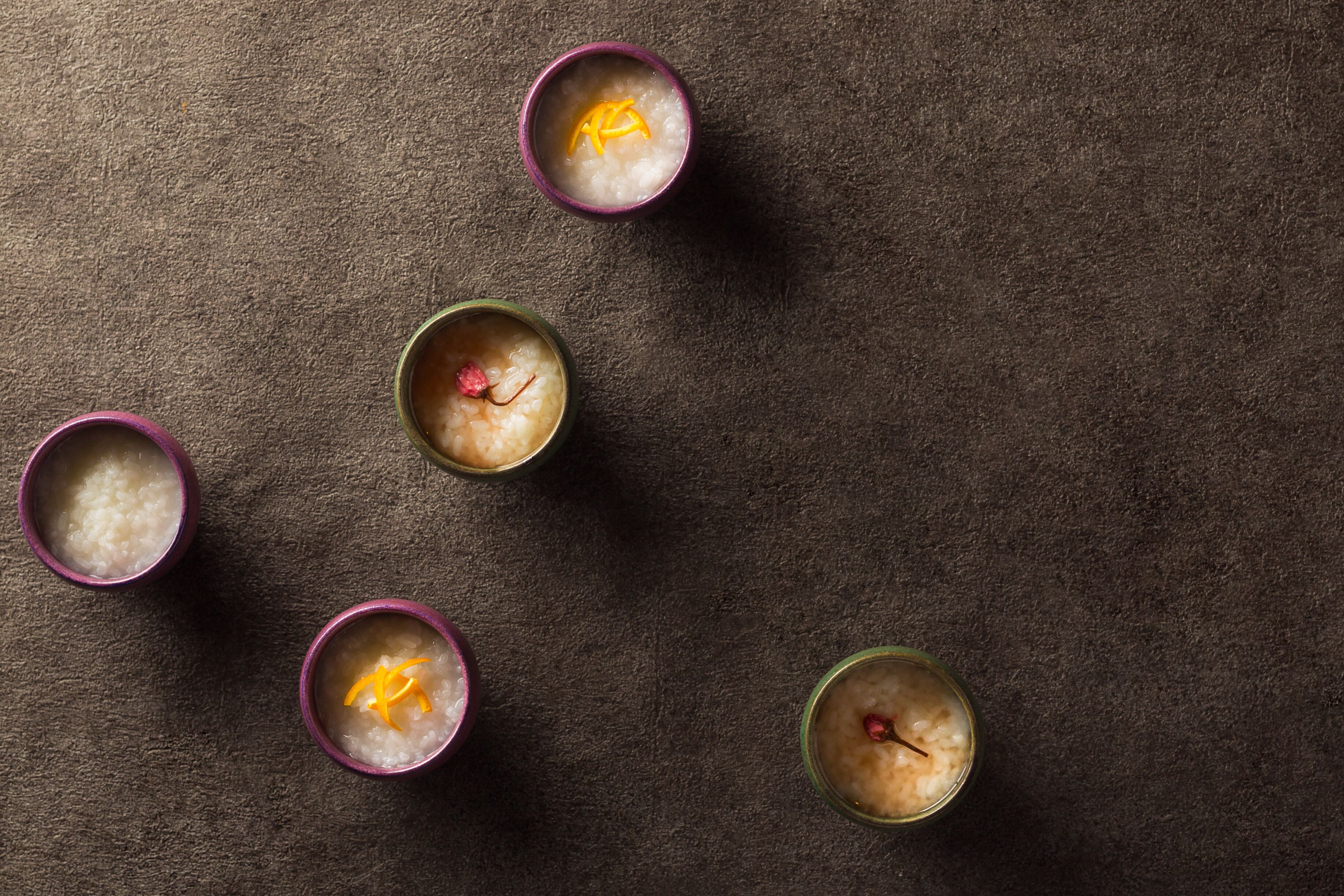 Five bowls of a white, grainy substance with a pinkish hue, each containing a small red object and a yellow peel, arranged on a dark textured surface.
