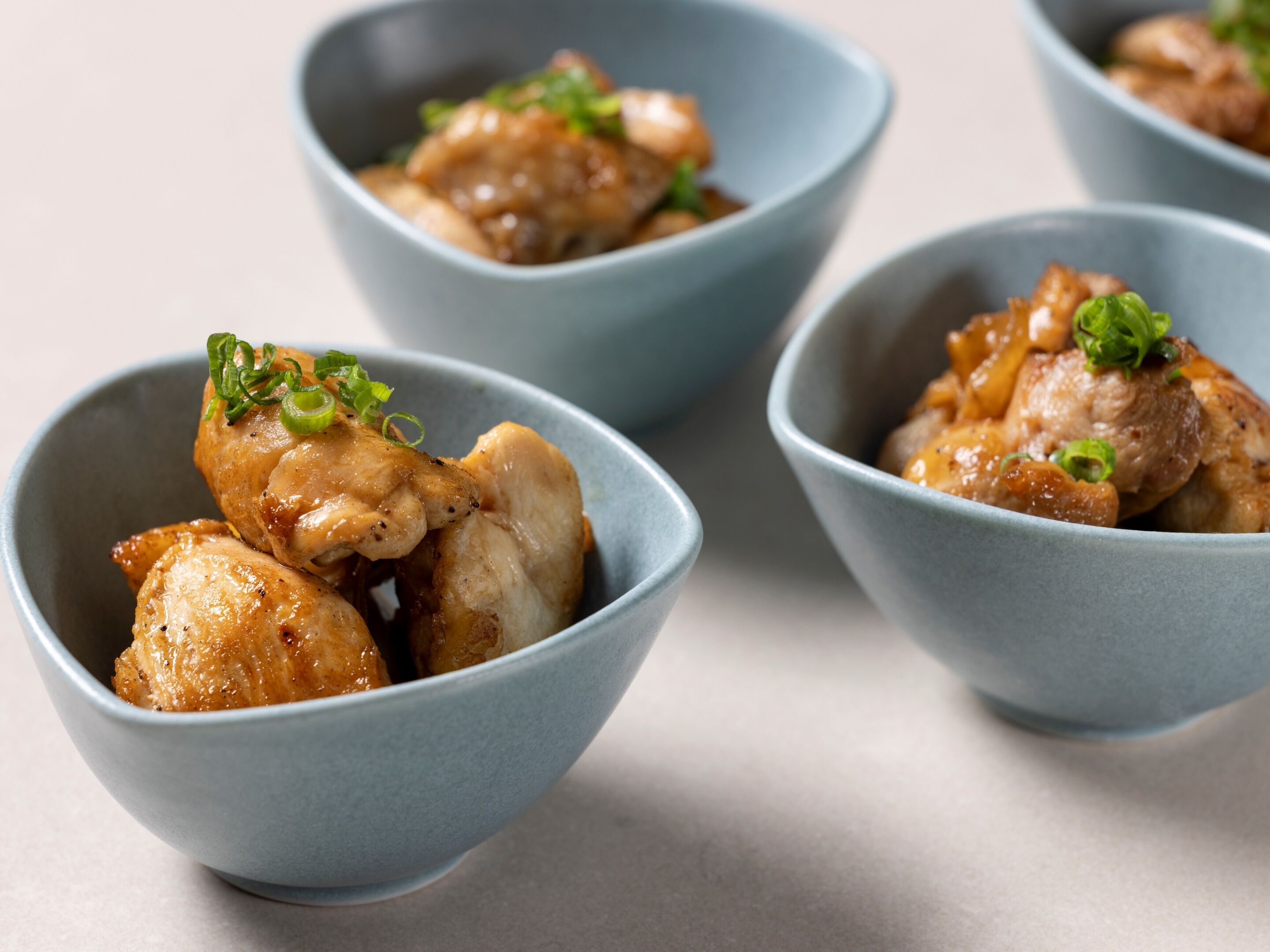 Four bowls of food with a light blue interior, containing pieces of chicken and garnished with green onions.
