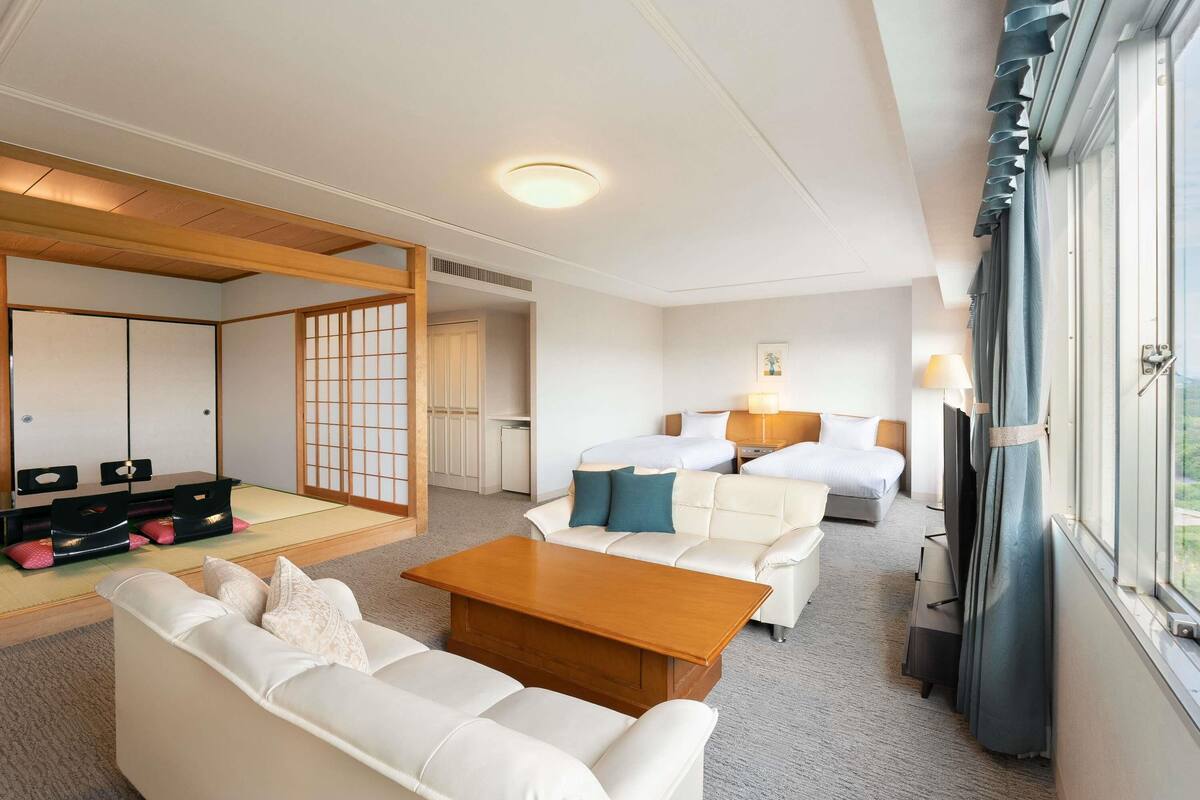 A hotel room with a traditional Japanese-style room divider, a sofa, a wooden coffee table, two beds with pillows, a nightstand with a lamp, and a view of the outdoors through a large window.