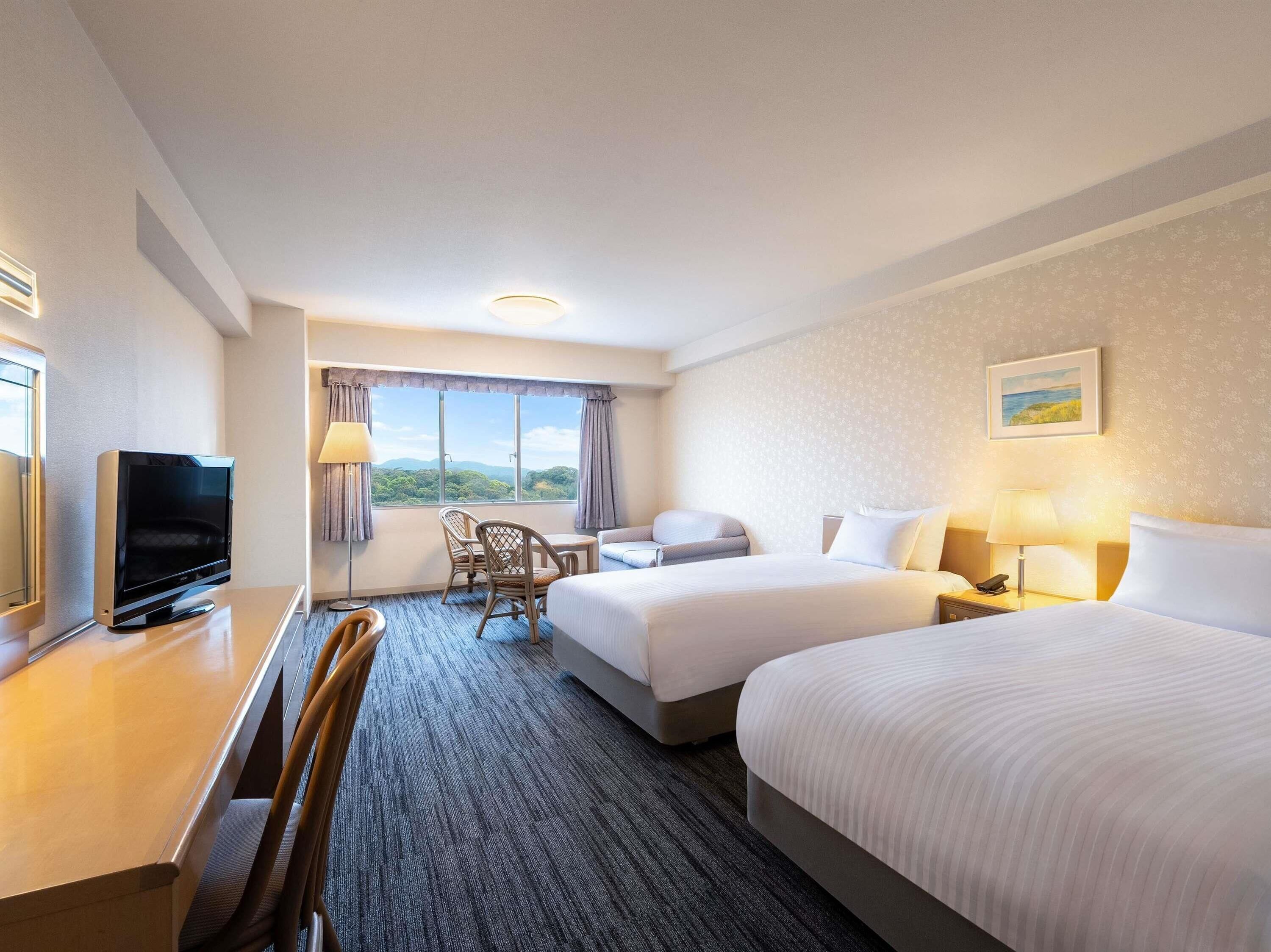 Ein Hotelzimmer mit zwei Betten, einem Schreibtisch mit Fernseher, einem Sessel, einer Schreibtischlampe und einem Fenster mit Blick.