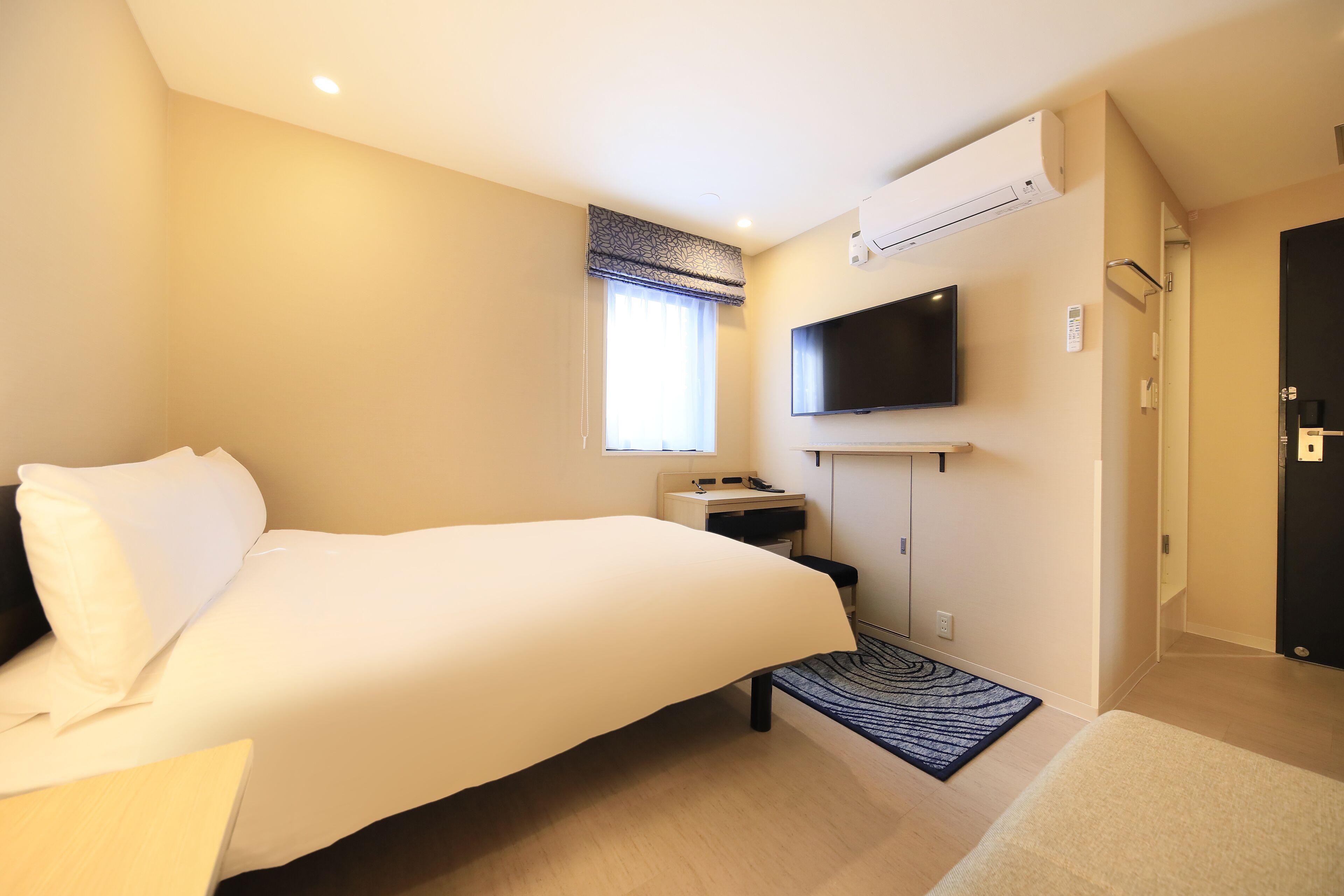 A hotel room with a bed, a desk, a flat-screen TV mounted on the wall, and an air conditioning unit.