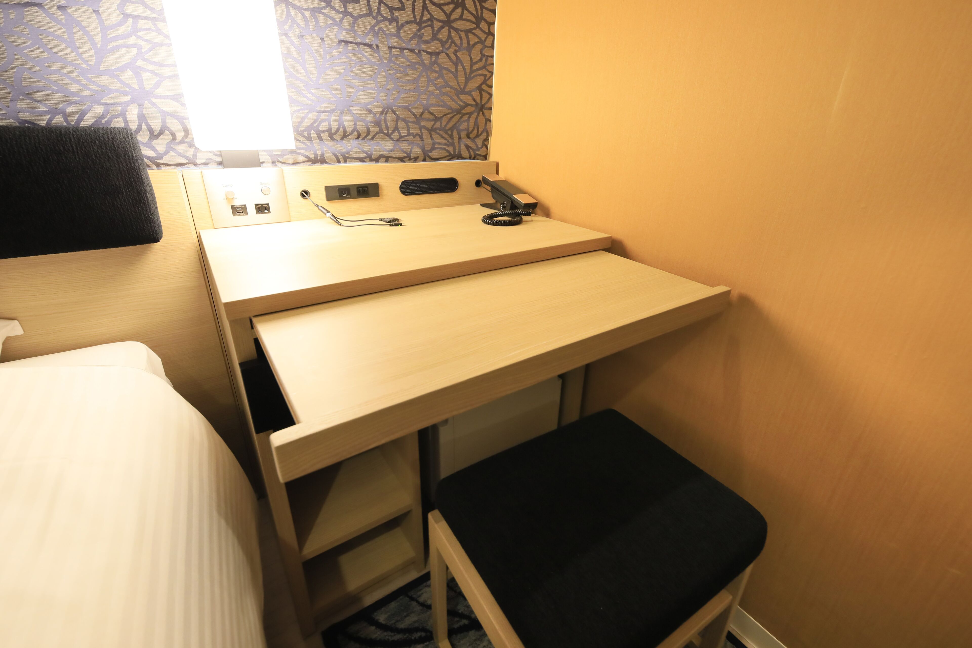 A compact room with a bed, a desk with a chair, a small stool, and a window with patterned curtains.