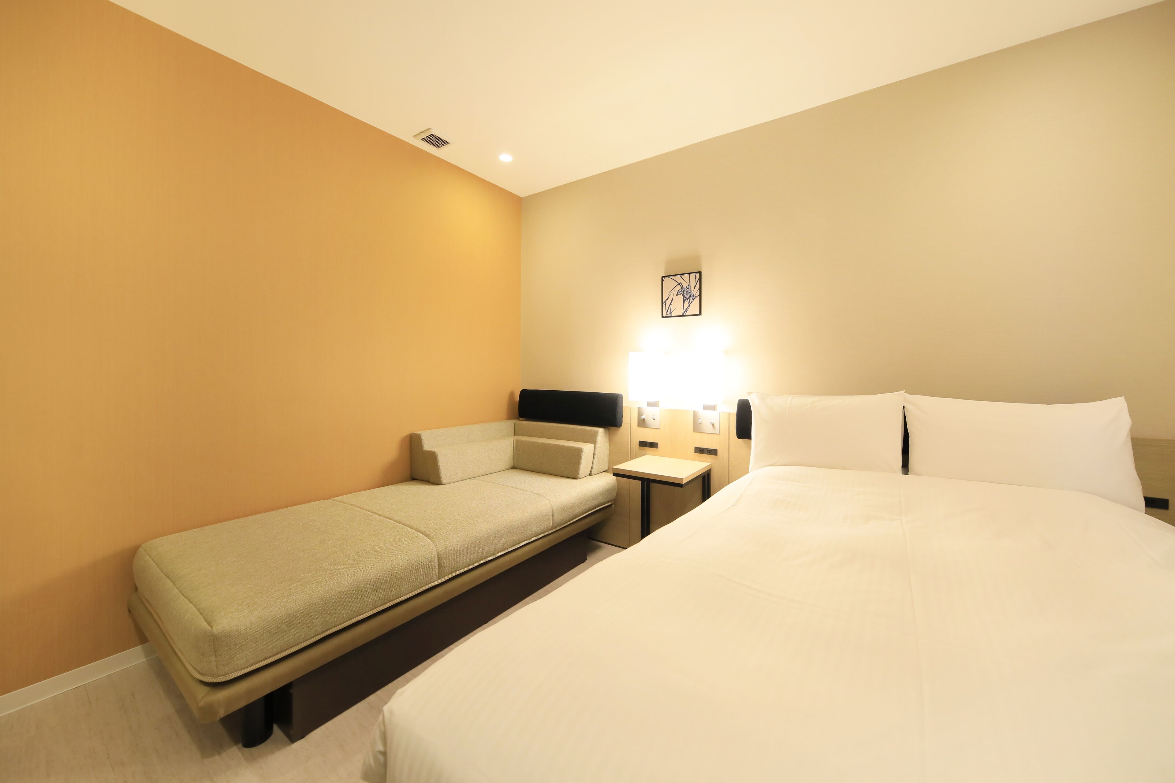 A hotel room with a large bed, a smaller sofa bed, a bedside table, and a wall-mounted light.