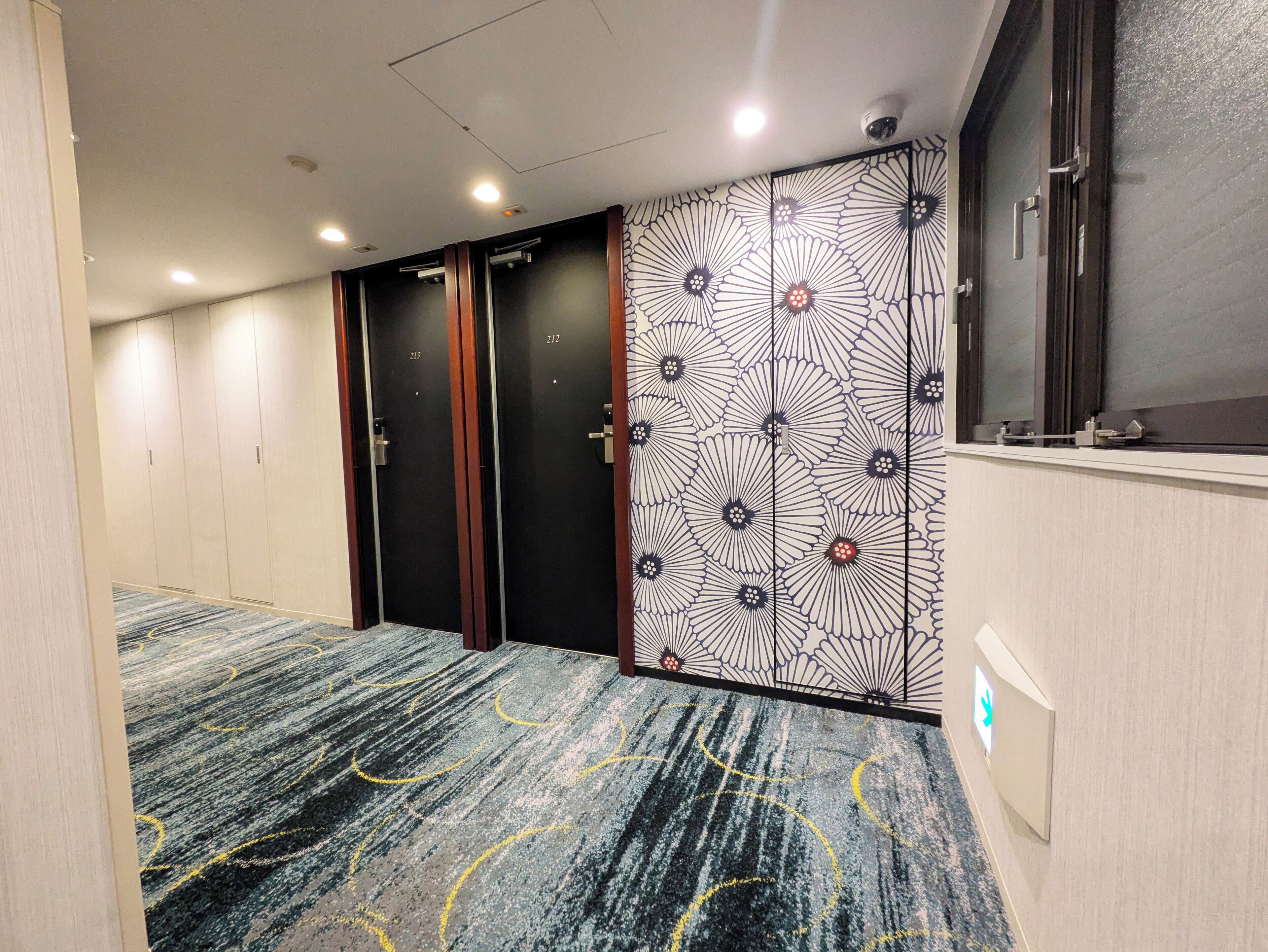 Un pasillo de hotel con alfombra estampada, una pared con diseño floral y una puerta con número.