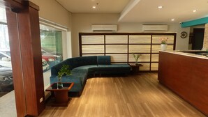 A reception area with a wooden reception desk, a green sectional sofa, a small wooden table with a plant, and a large window with a view of parked cars. - El Cielito Hotel (Makati)