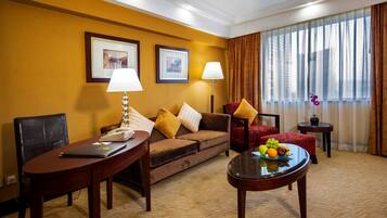 A hotel room with a brown sofa, two side tables, a small round table with fruit, and a large window with curtains.