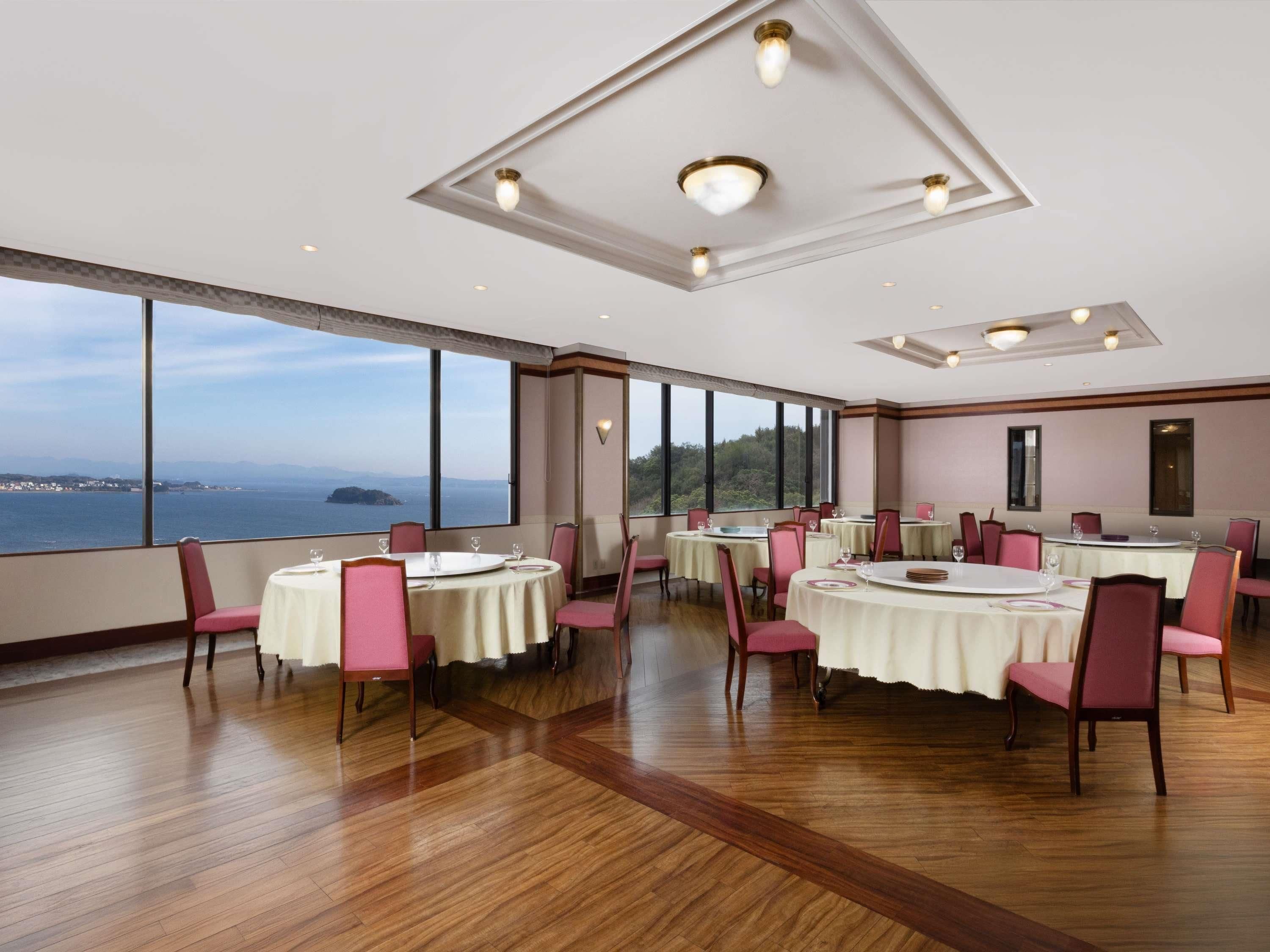 A dining area with round tables, chairs, and large windows offering a view of the sea.