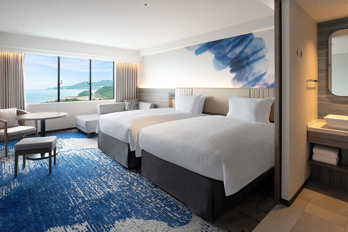 A hotel room with two beds, a seating area, a large window with a view, and a modern bathroom.