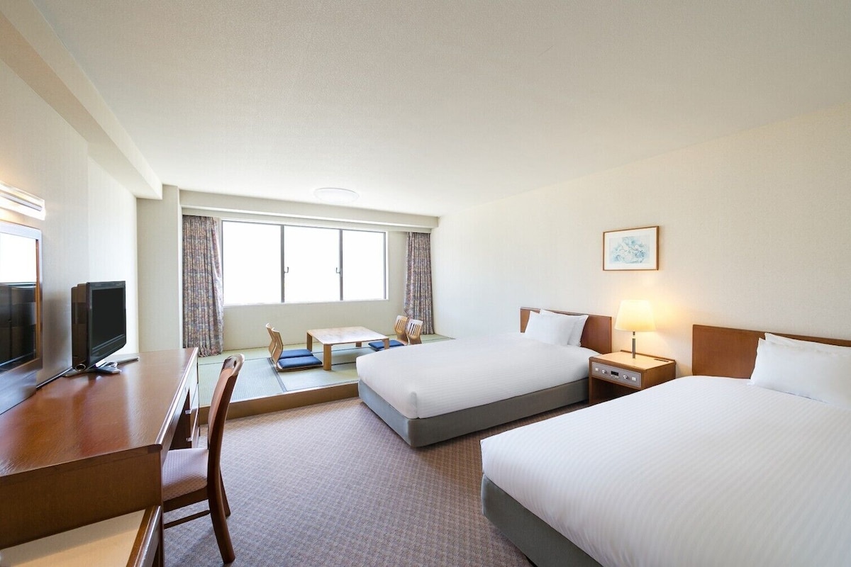 A hotel room with two beds, a desk, a TV, and a window with curtains.