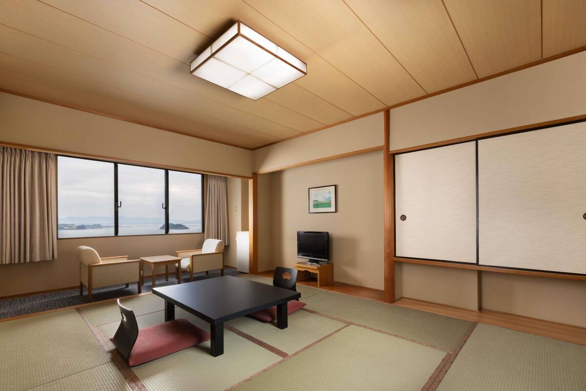 Una habitación con suelo de tatami, televisión, escritorio, silla, sofá y ventana con vistas, además de una puerta corredera.