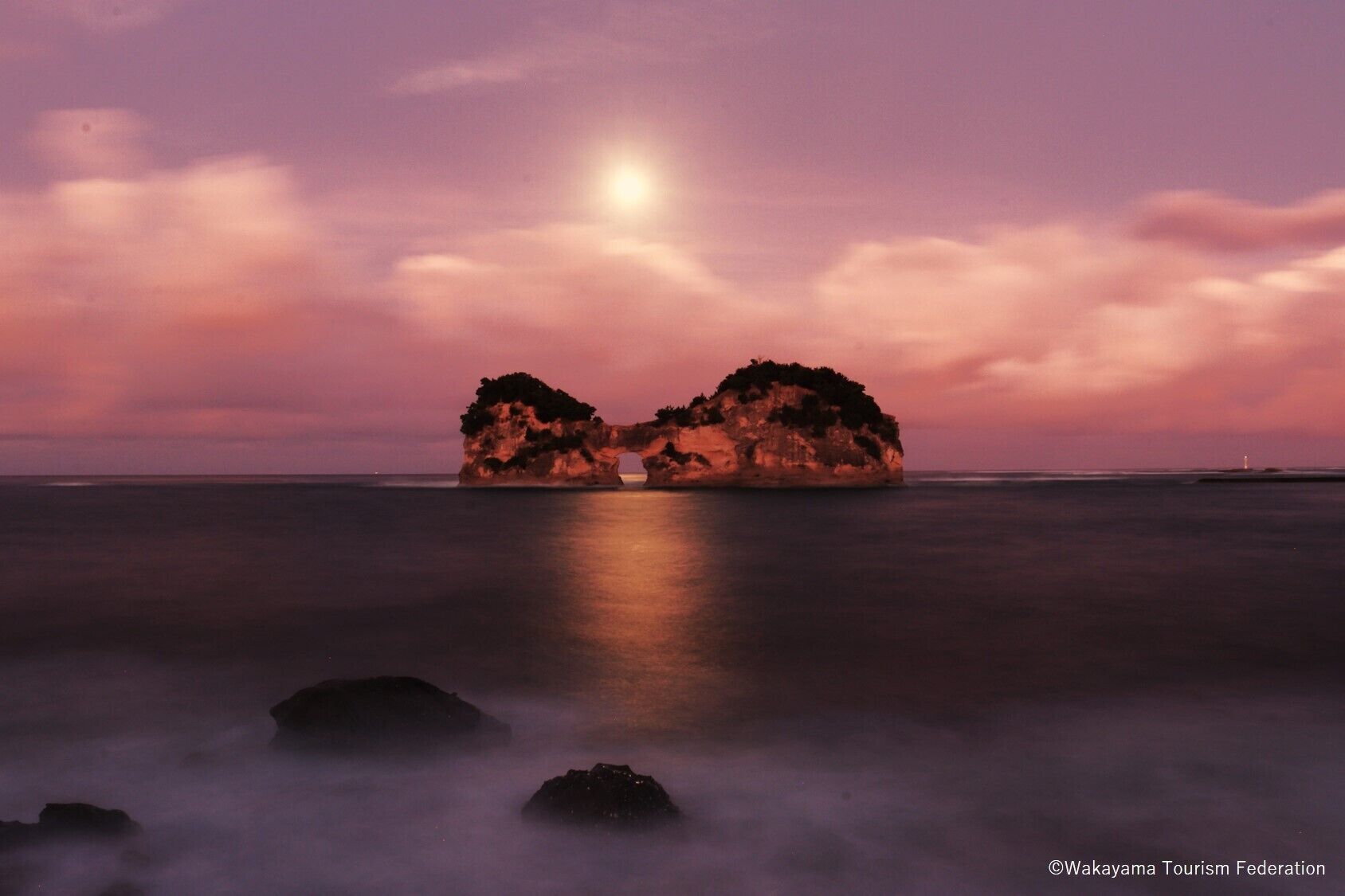 A calm sea at sunset with two islands and a lighthouse in the distance.