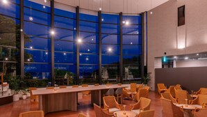 Un ambiente interno moderno con un lungo tavolo e sedie in vimini, ampie finestre e vista all'esterno.