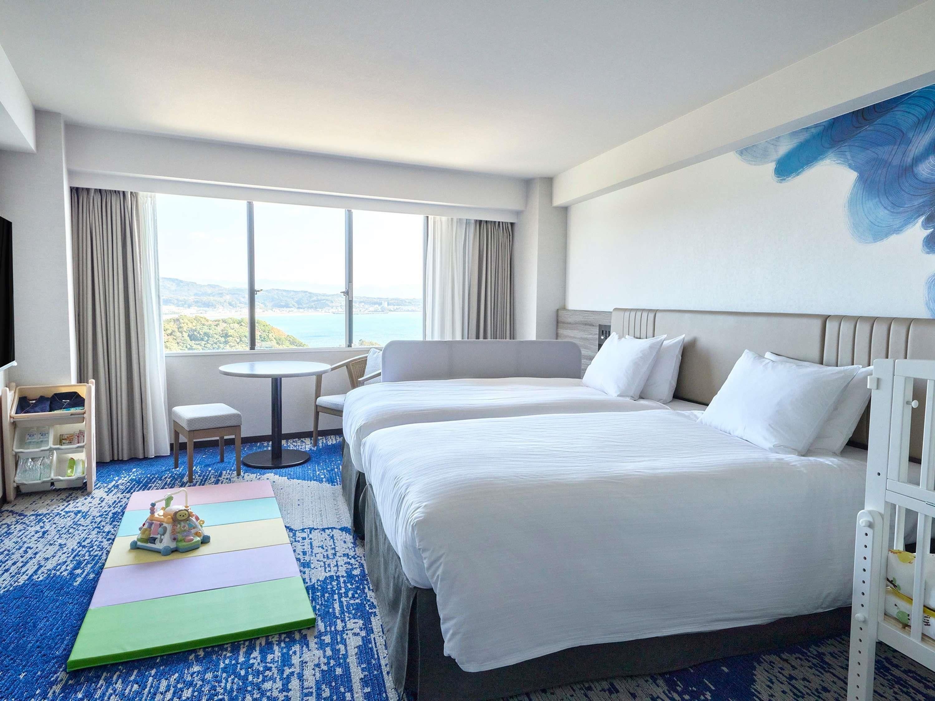 Habitación de hotel con una cama grande, una mesita, una silla, un tapete de juegos colorido y vista al mar.