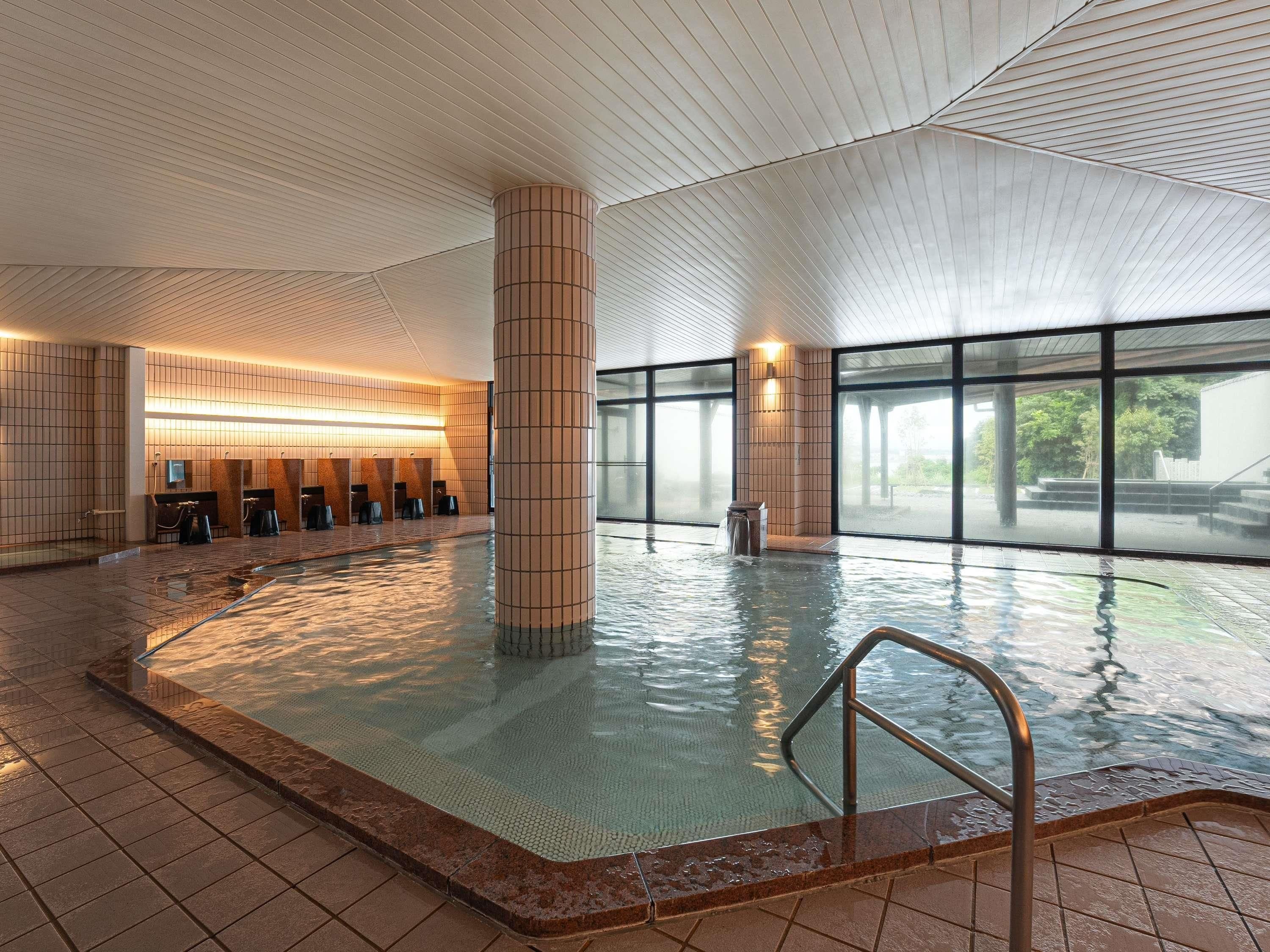A large indoor swimming pool with tiled walls and a central column, surrounded by windows.