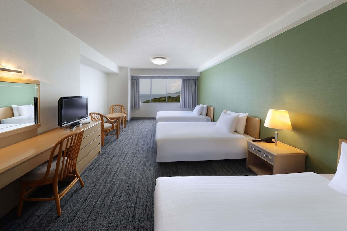 A hotel room with two beds, a desk with a TV, a chair, a lamp, and a window with a view.