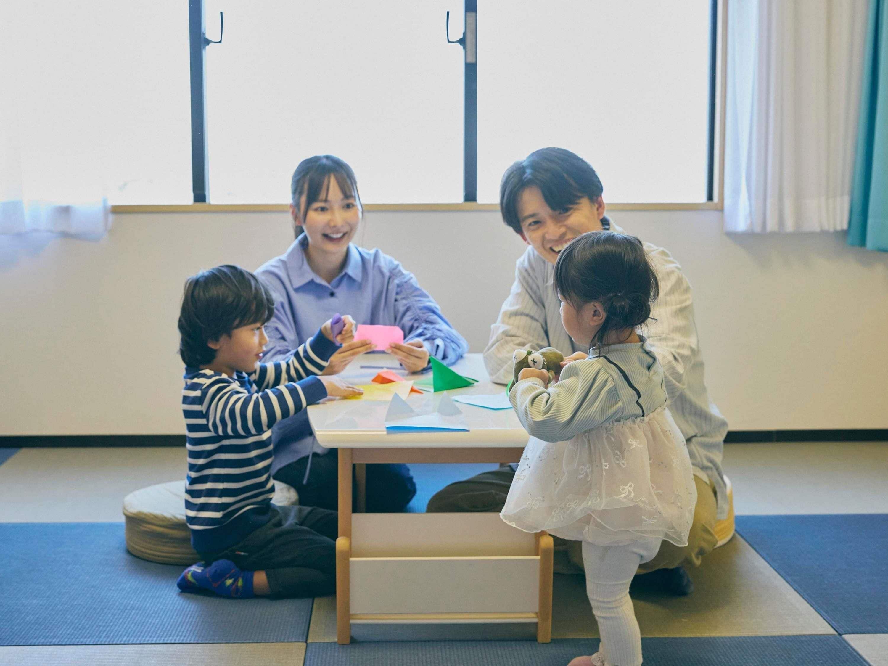 A group of children and an adult sitting on tatami mats around a low wooden table in a room with tatami flooring and a large window.
