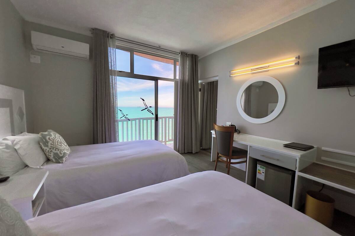 A hotel room with two beds, a desk, a chair, a TV, and a balcony with a view of the sea.