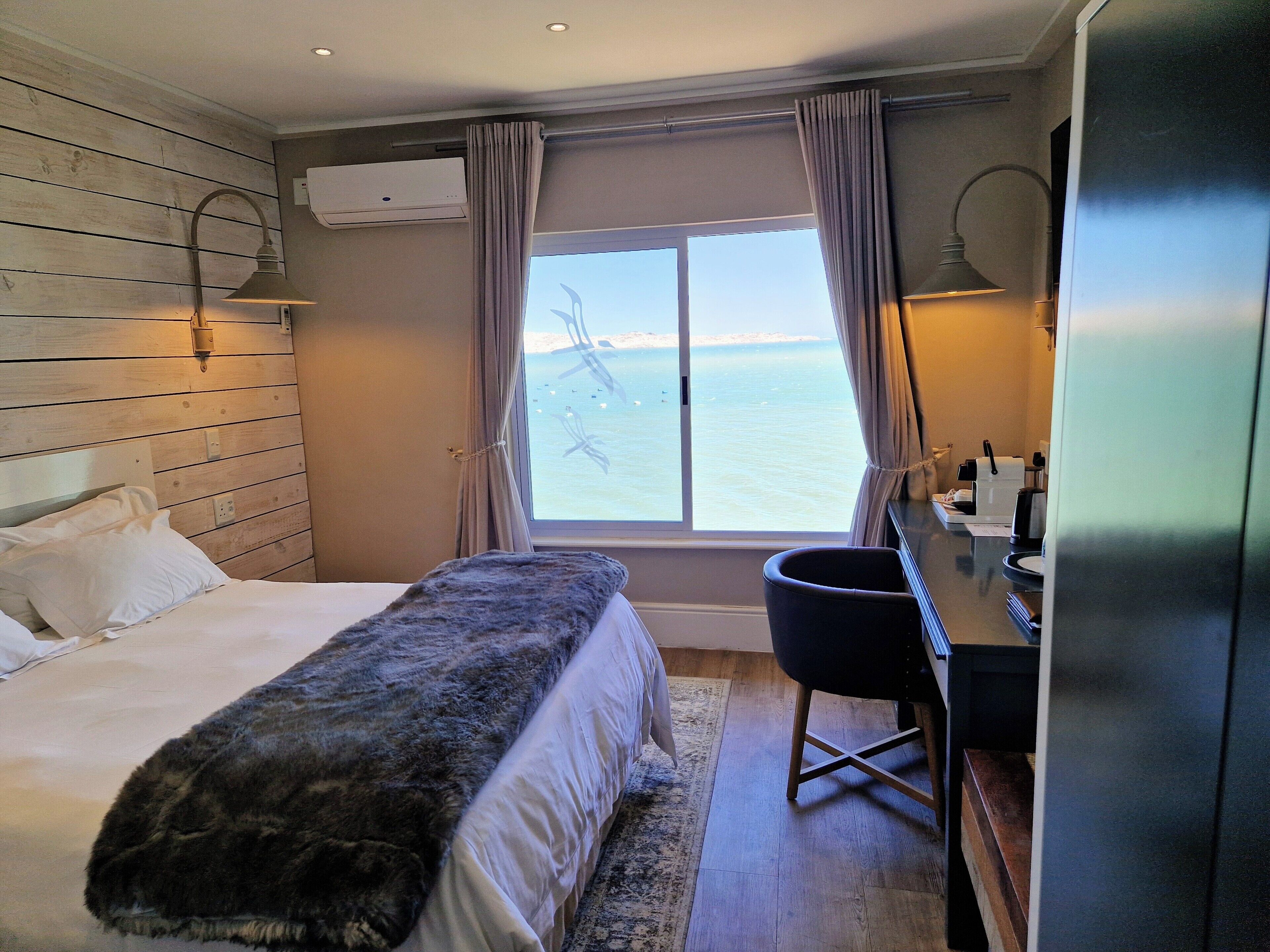 A hotel room with a bed, a desk, a chair, a window with a view of the sea, and an air conditioning unit.