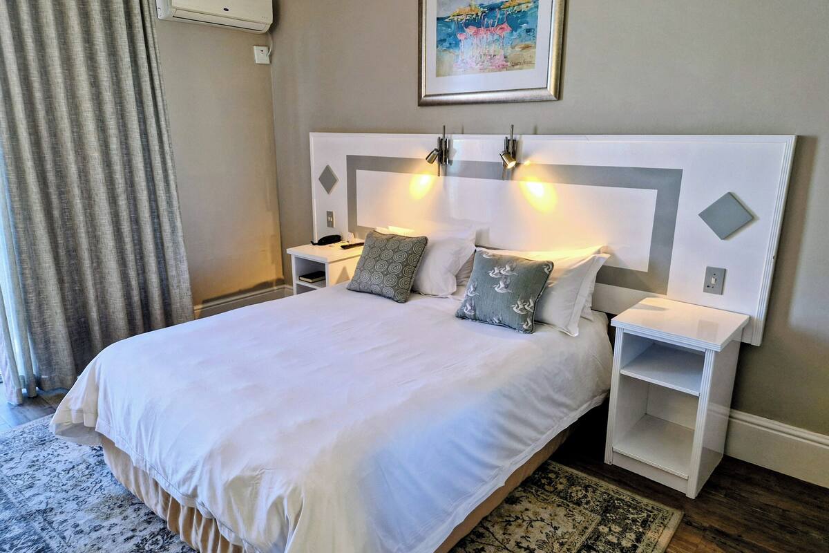 A neatly made bed with white linens, two bedside tables with lamps, a painting on the wall, and a patterned rug on the floor.