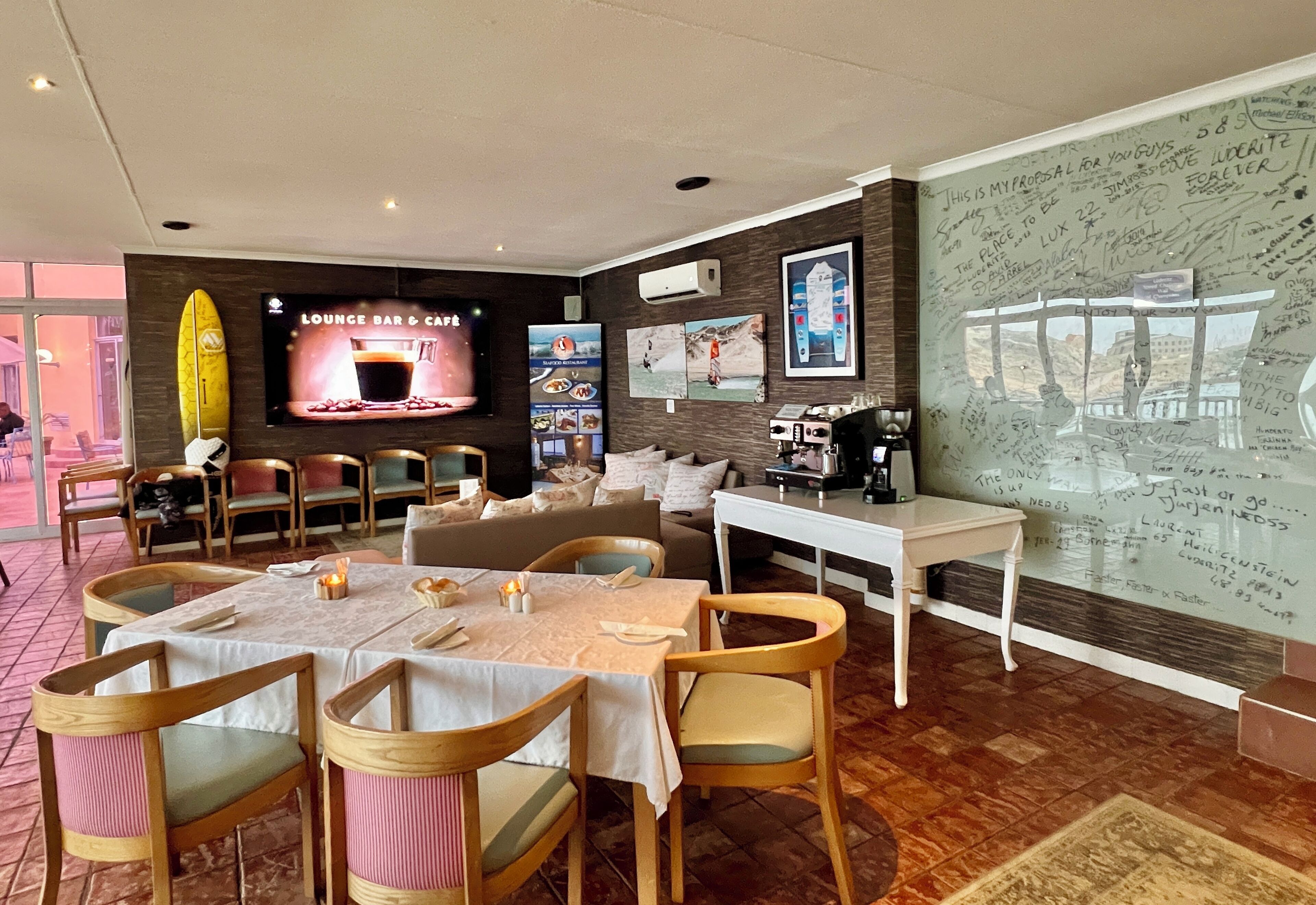 Un restaurant doté d’un écran de télévision affichant le texte « Lounge Bar & Café », de chaises en bois, d’une table blanche et d’un mur recouvert de notes manuscrites.