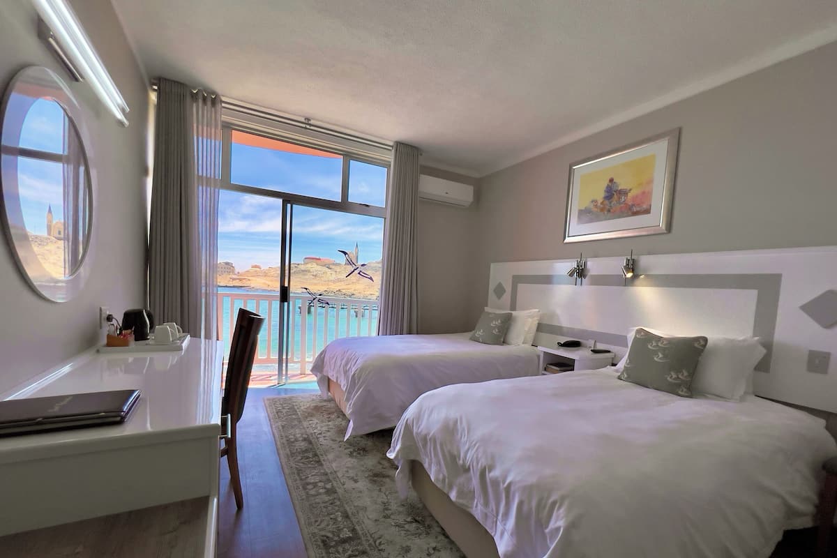 A hotel room with two beds, a desk, a chair, a large window with a view of the sea, and a painting on the wall.
