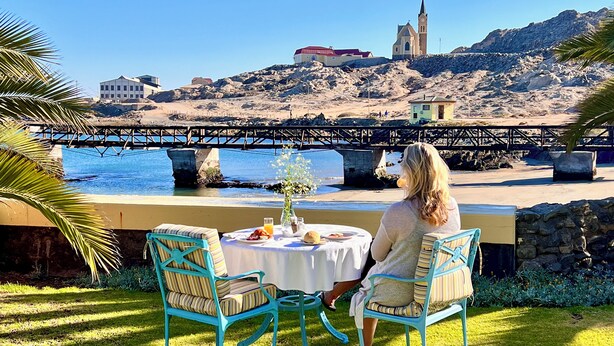 Una mesa con un jarrón de flores, un vaso de zumo de naranja y platos de comida, con vistas a una playa que tiene un puente y edificios al fondo.