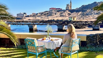 Una mesa con un jarrón de flores, un vaso de zumo de naranja y platos de comida, con vistas a una playa que tiene un puente y edificios al fondo.