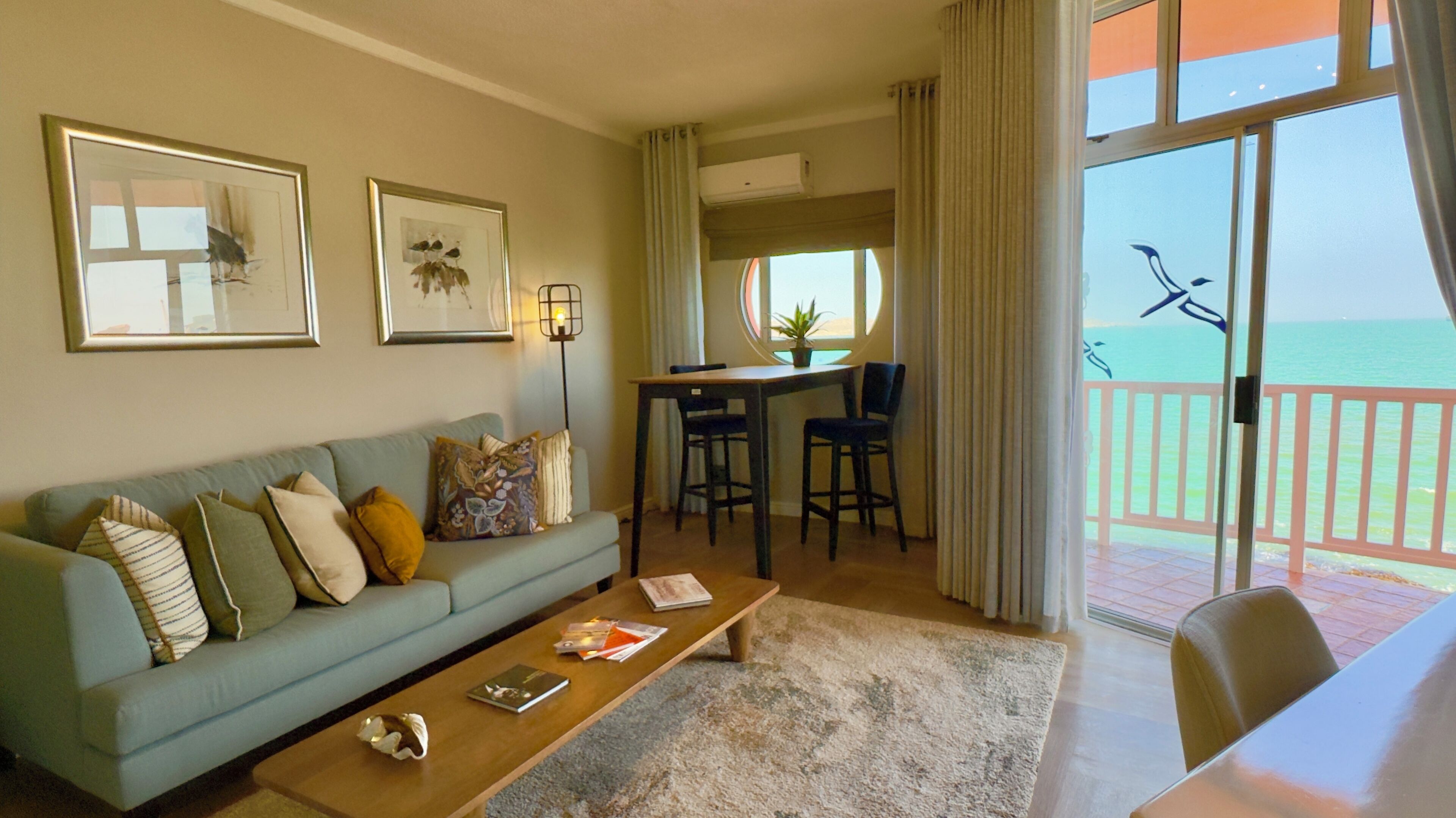 A living room with a sofa, coffee table, and artwork on the wall. There is a balcony with a view of the ocean.