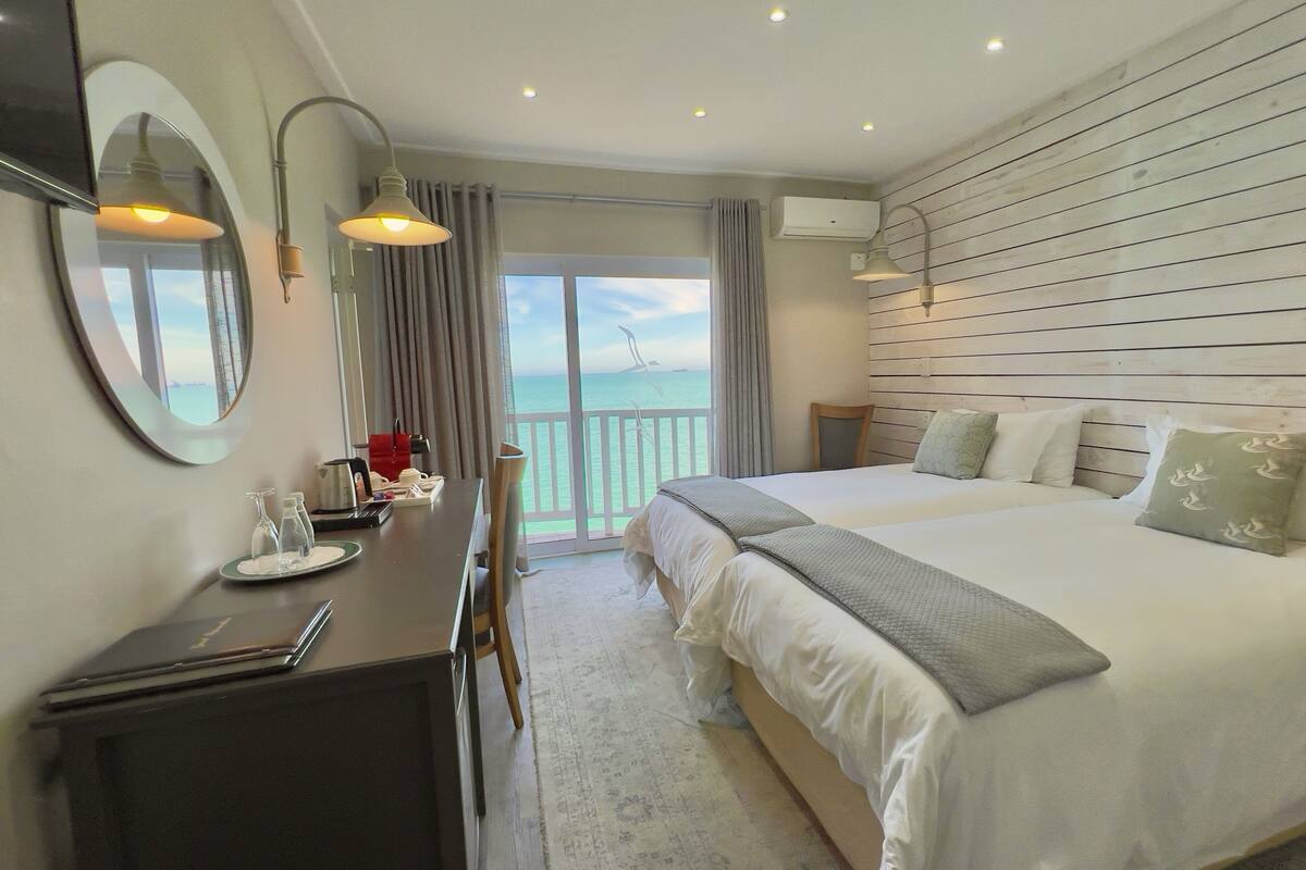 A hotel room with a large bed, a desk with a laptop, a chair, and a view of the sea through a sliding door.