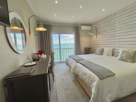 Habitación de hotel con una cama grande, un escritorio con una computadora portátil, una silla y vista al mar a través de una puerta corrediza.