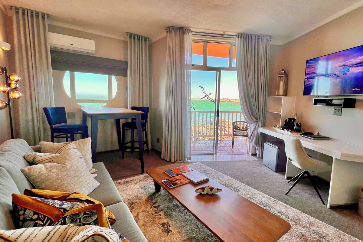 A modern hotel room with a sofa, a dining table with chairs, a desk with a computer, a TV, and a view of the sea through a glass door.