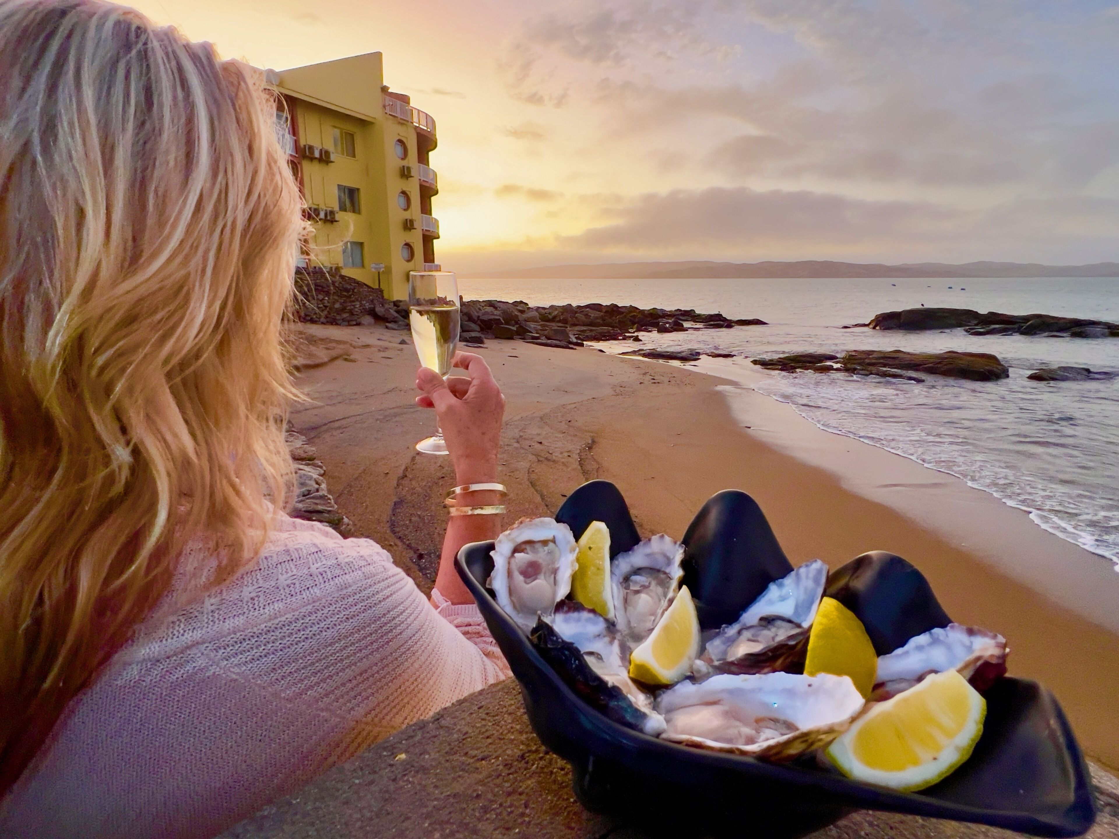 Une personne tenant un verre de champagne et un bol d’huîtres garni de tranches de citron, sur une plage, avec un bâtiment visible en arrière-plan.