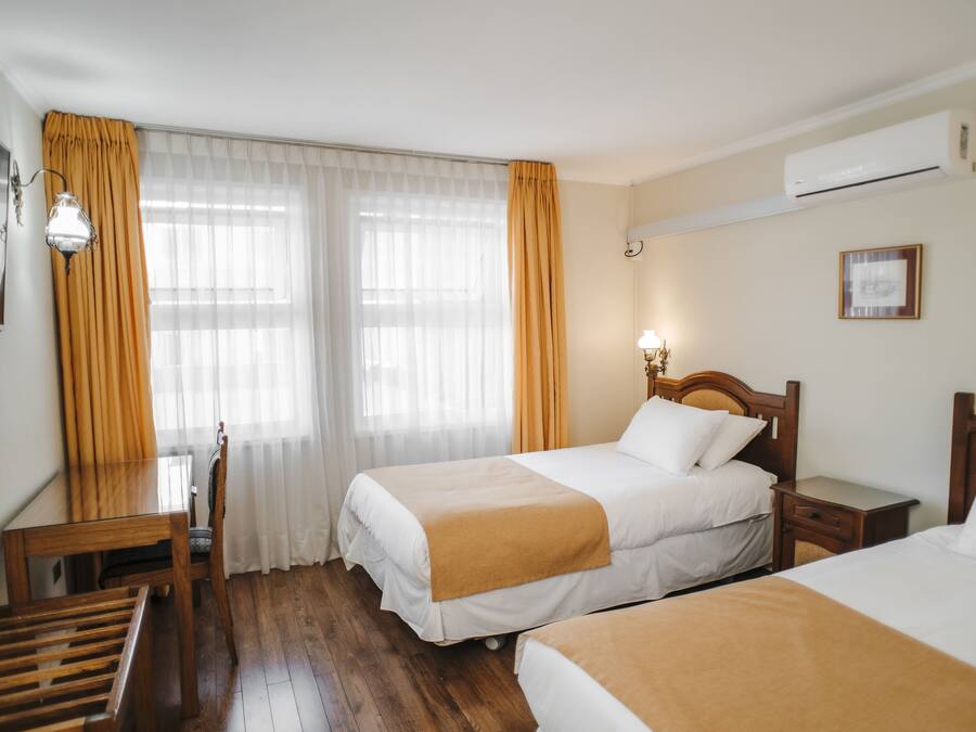 Habitación de hotel con dos camas, un escritorio de madera, una silla y un ventanal con cortinas.